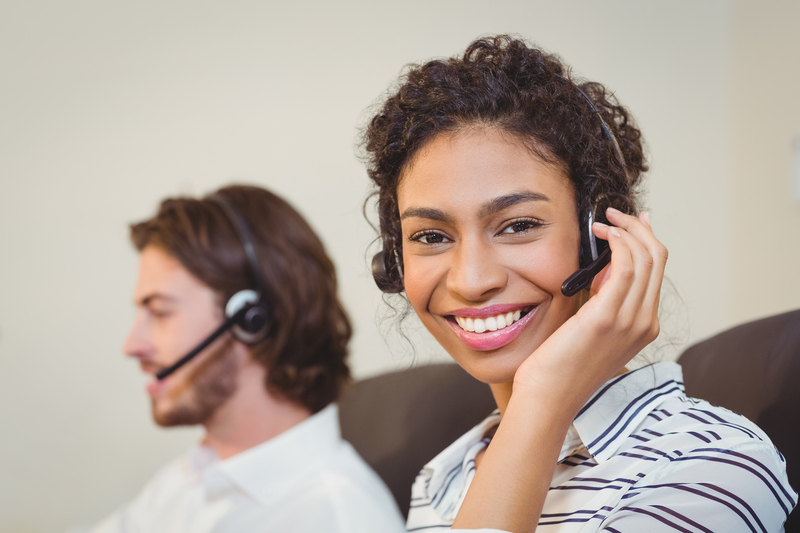 Bild på två kollegor i headset, som på helpdesk eller kundtjänst. Ena kollegan kollar in i kameran och ler med handen på öronsnäckan.