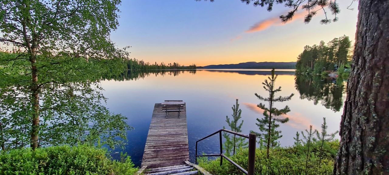 Brygga vid en spegelblank sjö, med lite växtlighet på sidan. Blå himmel. 