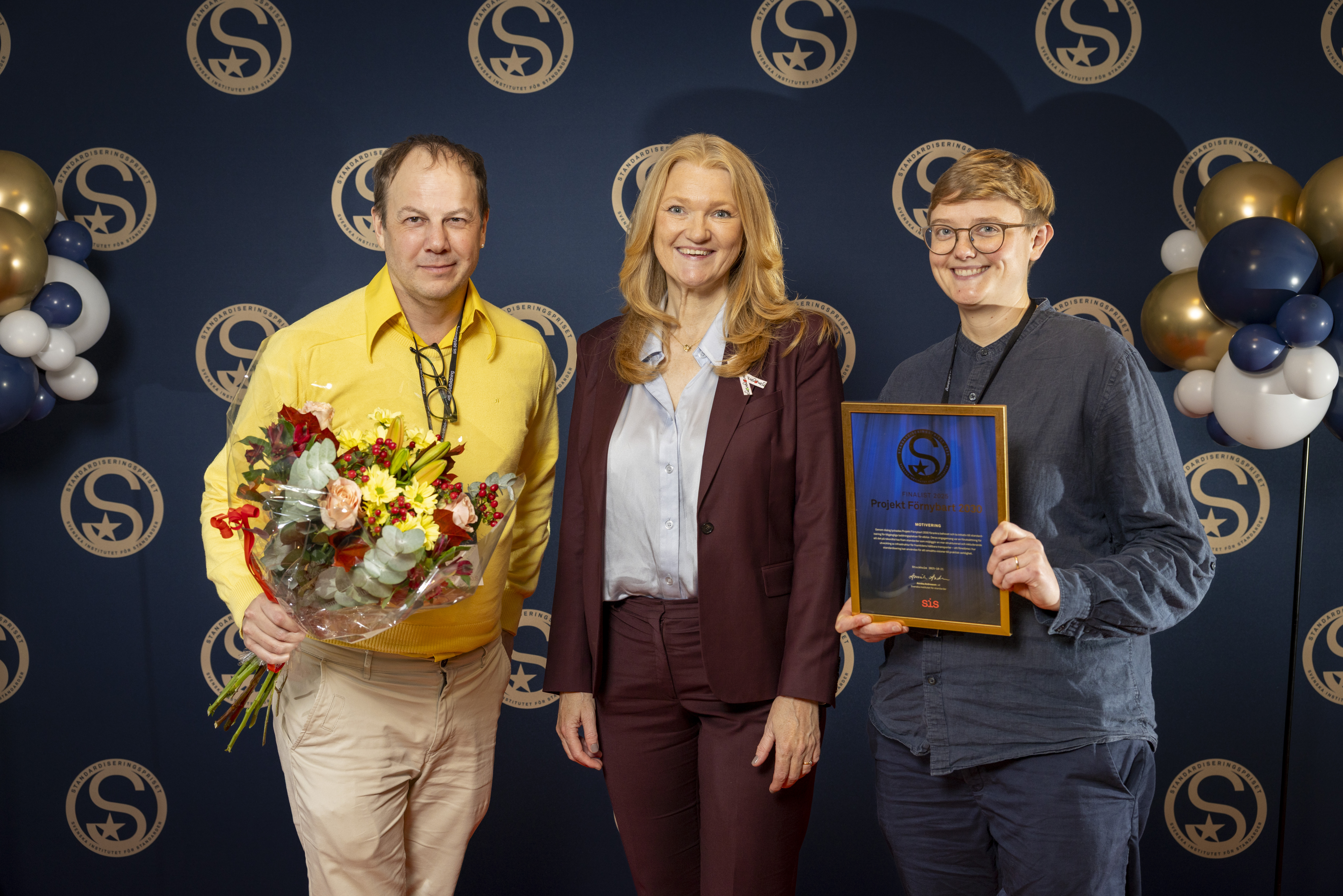 Tre personer står bredvid varandra omgivna av ballonger. Personen till vänster håller i en blombukett, den till höger håller ett inramat diplom.