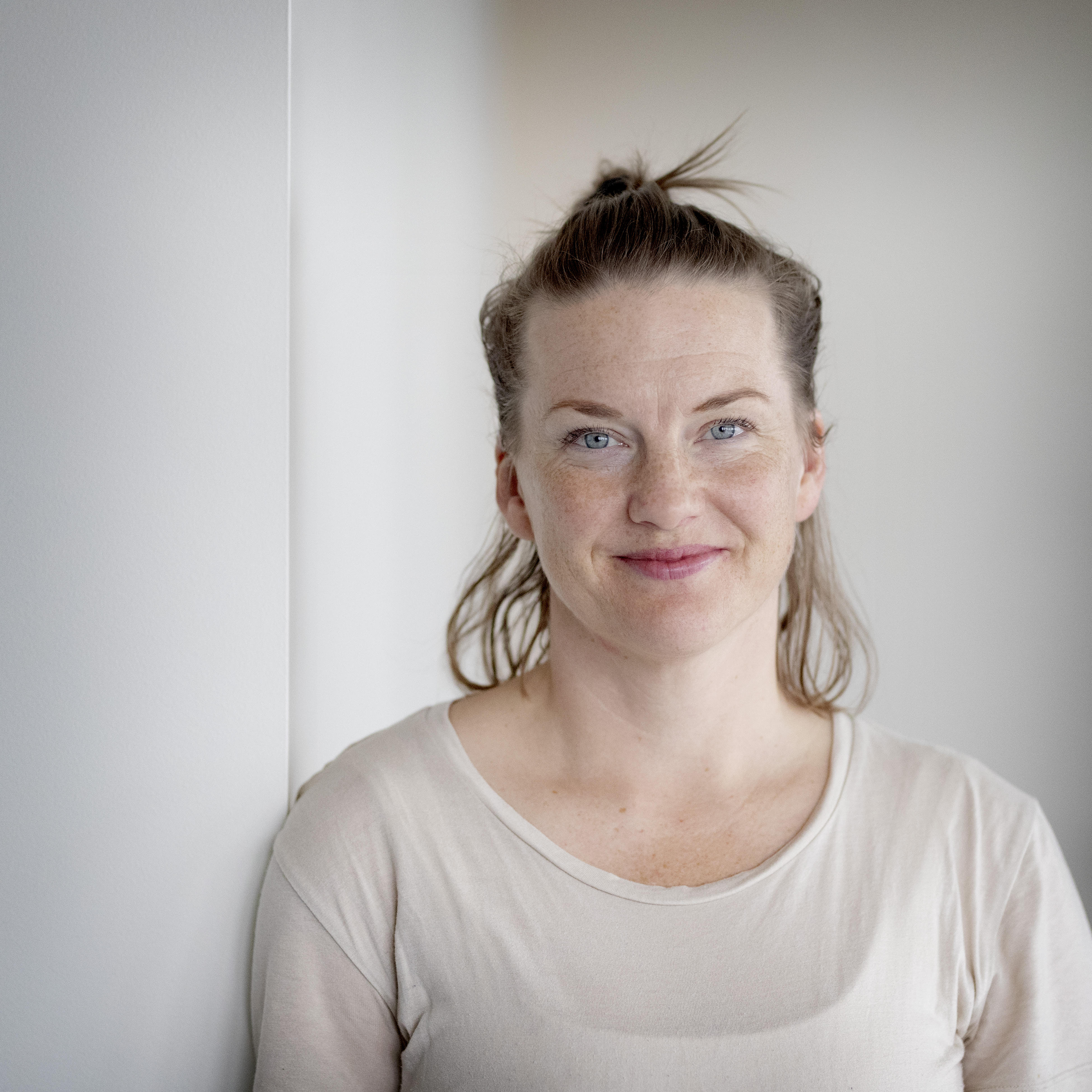 Ljus porträttbild på en mjukt leende person i ljusa kläder vid en vit vägg.