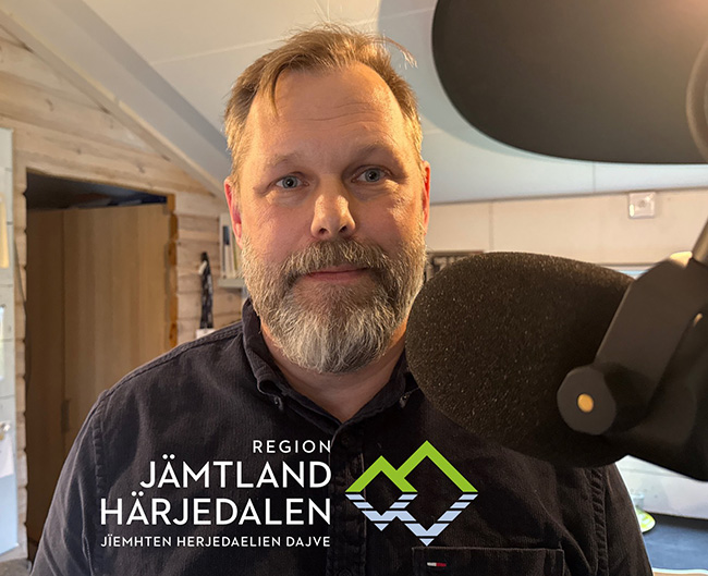 En man med grått hår, skägg och mustasch står i en studio framför en mikrofon