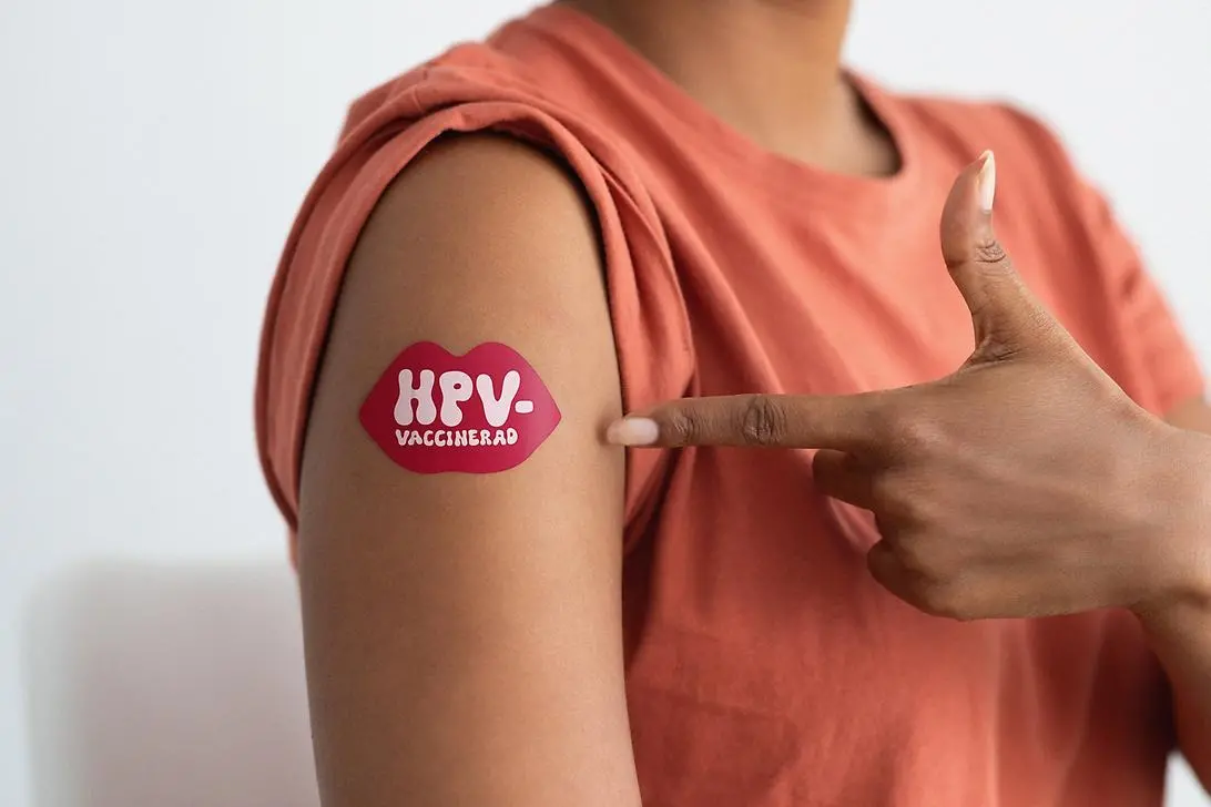 Person i halvbild pekar på sin högra överarm där det sitter ett plåster som är format som en röd mun vari det står HPV-vaccinerad.