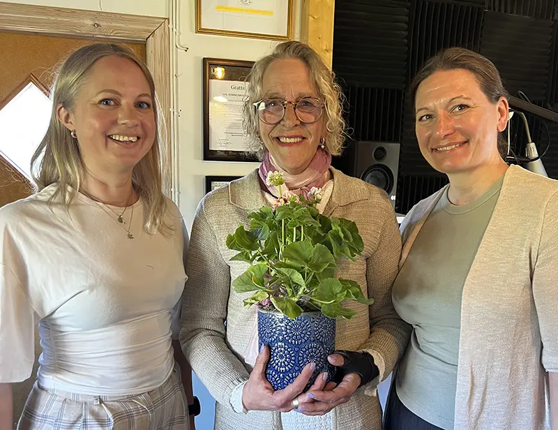 Tre kvinnor står uppradade och ler in i kameran, den mittersta med en blomma i famnen.