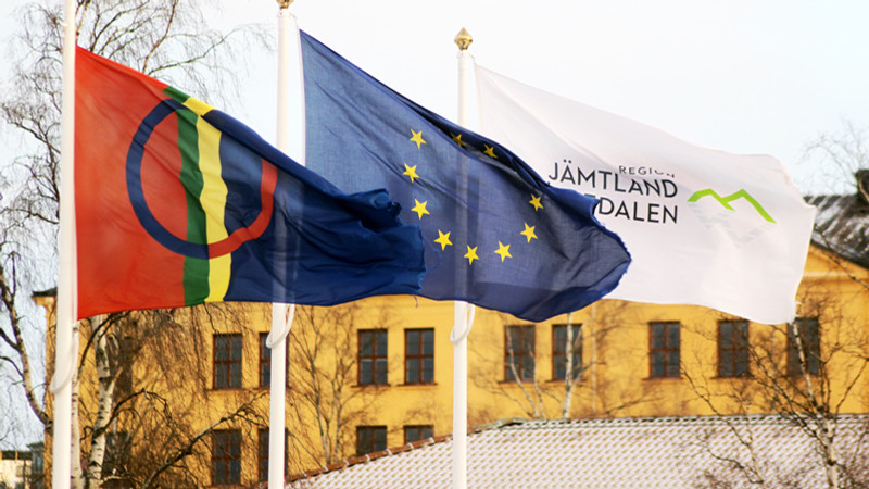 Tre flaggor som vajar i vinden: samiska flaggan, EU:s flagga och en flagga med Region Jämtland Härjedalens logotyp på.