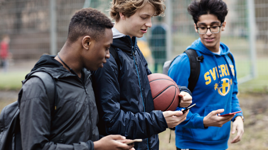 Tre vårklädda ungdomar går till eller från en träning med basketbollar och mobiltelefoner i händerna.