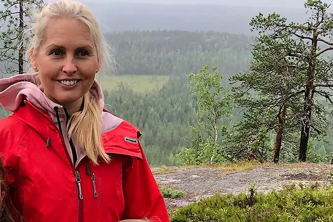 En ljushårig person i röd vindjacka ler mot kameran uppe i fjälltrakterna