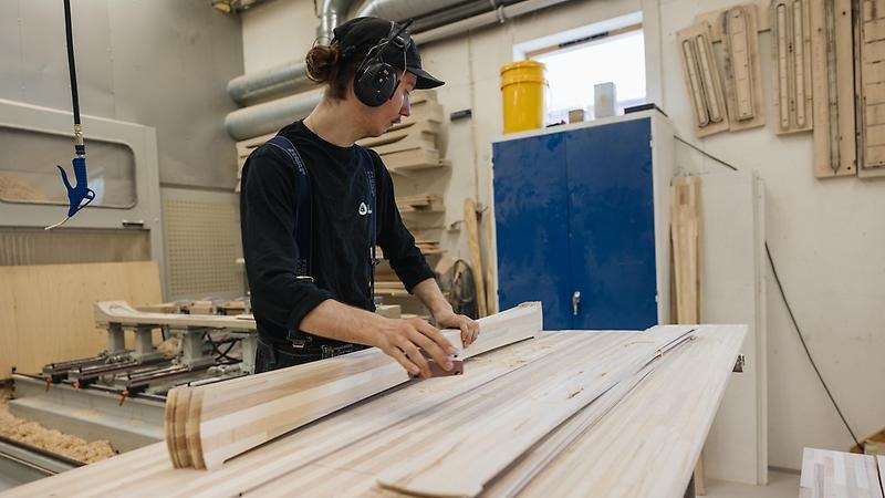 Fabriksarbetare står vid arbetsbänk med ett gäng träplankor i handen som ska bli till skidor.