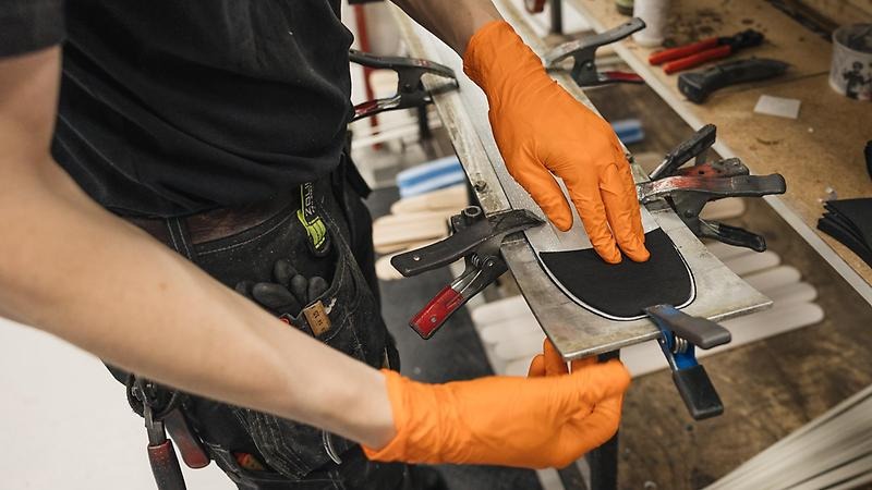 Närbild på ett par händer som jobbar med en skidspets som satts fast med tvingar och klämmor vid en arbetsbänk.