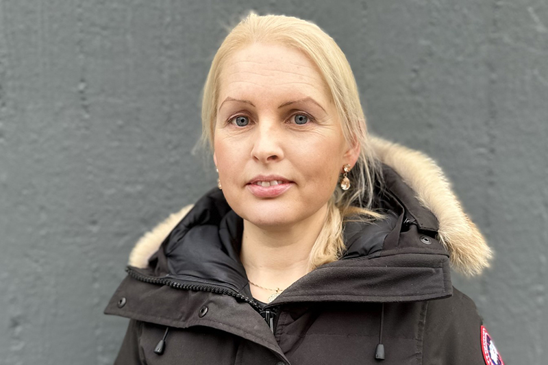 En person i vinterjacka med blont hår uppsatt i en hästsvans tittar in i kameran