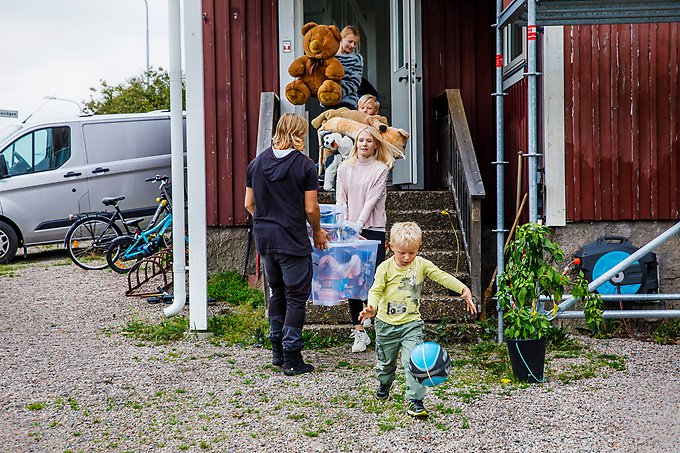 Två vuxna och tre barn står utanför ett rött hus med vita knutar. De vuxna och två barn står i trappan till huset och bär på stora platslådor och gosedjur. Ett barn leker med en boll nedanför trappan. Till vänster om huset syns två cyklar och en grå skåpbil.