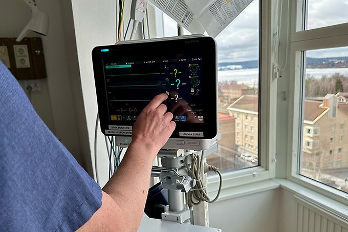 En arm kommer upp i neder vänsterkant av bilden och trycker på en skärm med patientdata. Skämen står invid ett fönster med utsikt från höghuset på sjukhuset.