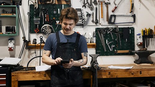 Verkstadsarbetare står i förkläde vid sin arbetsbänk med verktyg, och gör något på pekskärmen på sin mobiltelefon.
