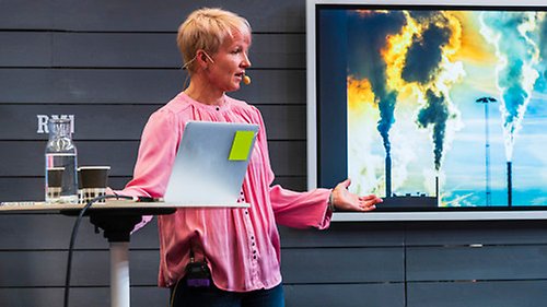 Föreläsare på scen under mässa, presenterar med en skärm bakom sig. Bilden på skärmen visar industriskorstenar med rök.