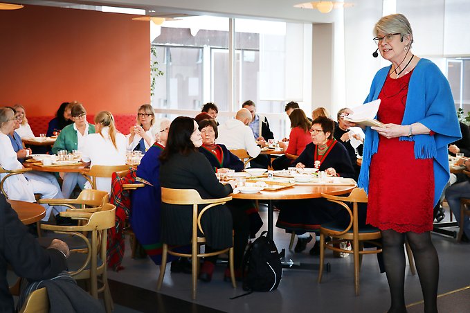 en kvinna klädd i röd klänning och blå sjal håller tal inför en publik som sitter ner och äter lunch