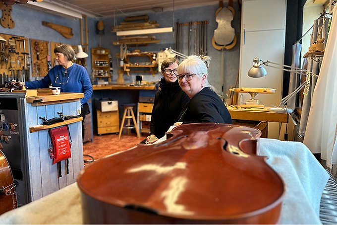 Interiör från Astrids fiolverkstad med en liggande kontrabas i framkant. I bakgrunden syns tre kvinnor