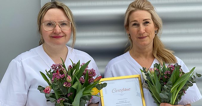 Två vitklädda personer står utomhus mot en vägg med blombukett och diplom i sina händer